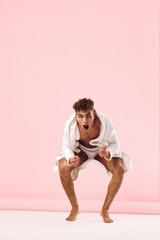 Excited young man in white bathrobe, striking a playful pose against a pink background, showcasing...