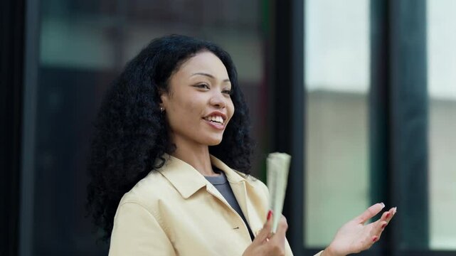 Cheerful woman holding a stack of cash while gesturing with confidence, symbolizing financial success in an urban setting.