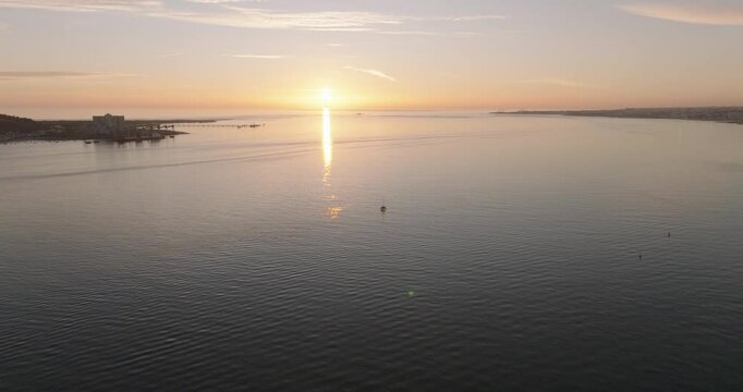 Aerial drone shot of the Tagus river, Atlantic Ocean and horizon at sunset in Lisbon, Portugal, Europe. Catamaran facing the sun. Forte do Bugio lighthouse visible. Shot in ProRes 422 HQ