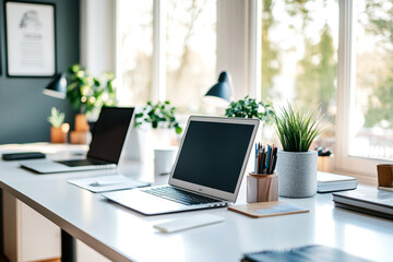 Fototapeta premium Bright and modern workspace with laptops, potted plants, stationery, and large windows letting in natural light