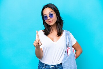 Fototapeta premium Young caucasian woman holding a beach bag isolated on blue background showing and lifting a finger
