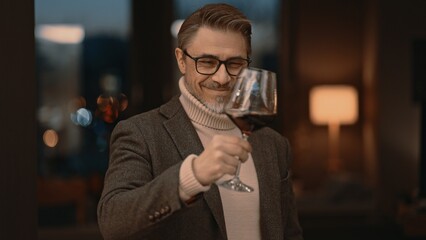 Elegant mid adult man drinking red wine from glass at home in cosy room. Portrait of happy, satisfied middle aged man, smiling. Satisfied wine expert tasting.  .