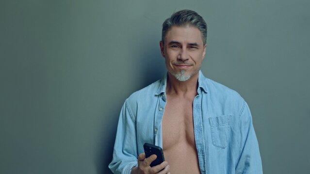 Confident mature man in casual posing with a smartphone against a gray backdrop in open shirt at morning. Mid adult male in 50s, bearded, grey hair, friendly and approachable, smiling.