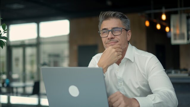 Confident businessman thinking and smiling over laptop in office - Powered by Adobe