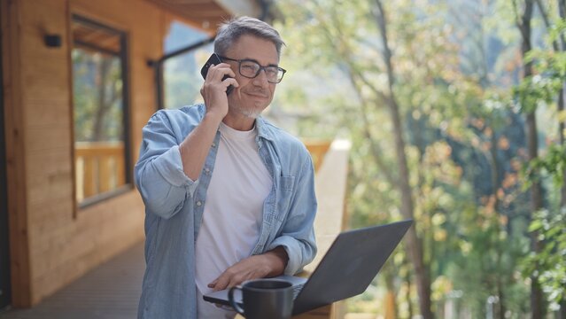 Mature freelancer working remotely on laptop and talking on phone on terrace of his wooden country house, enjoying fresh air and nature
