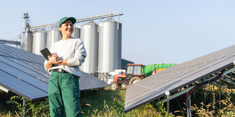 Female farmer with digital tablet on a modern farm using solar panels. Agricultural silos in the...