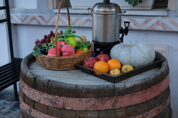 A rustic setup on a wooden barrel featuring a wicker basket filled with fresh fruits like apples, grapes, and pomegranates. Next to it, an assortment of pumpkins, oranges, and a traditional samovar, c