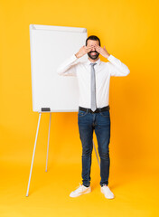 Full-length shot of businessman giving a presentation on white board over isolated yellow background covering eyes by hands