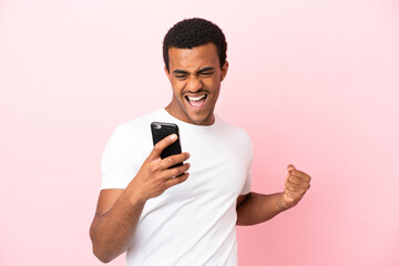 African American handsome man on isolated pink background using mobile phone and doing victory...