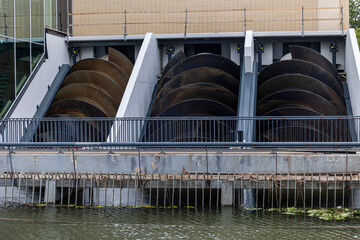 The image shows a set of large Archimedes screws used for water management or energy generation. The screws are housed within a structure beside a canal or river, with a metal railing in front.