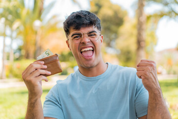Young hispanic man at outdoors holding wallet with money and celebrating a victory