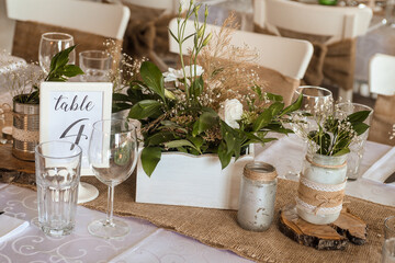 Wedding decoration on the table that is ready to welcome the wedding guests. The table is decorated with beige color, a bouquet of green and white flowers, as well as wooden materials and fabrics. It 