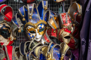 A close-up view of several ornate Venetian carnival masks. The masks are richly decorated with gold, blue, red, and black patterns. They hang on a wire rack, ready for purchase.