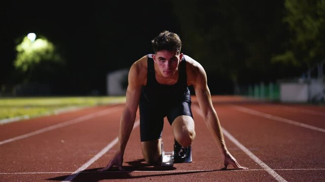 Strong Young Man Starting a Race From Track Starting Blocks Position on a Dark Stadium in the Evening. Cinematic Portrait of a Fit Male Sprint Runner Participating in a Competition