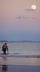 Gold prospector. Senior treasure hunter searching for jewelry with metal detector on beach with super moon on horizon. Vertical photo