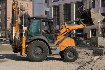 A yellow tractor repairs a road in the city with a bucket and tiles near the roadway during the day