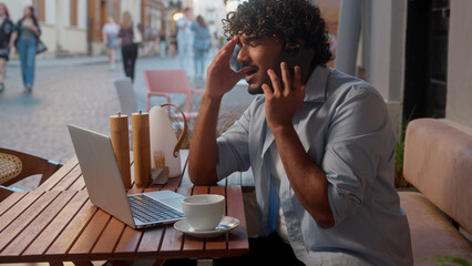 Indian Arabian man business guy businessman in cafe urban street online work laptop male tourist mobile phone call talk stress angry client problem cellphone conversation argue conflict city outdoors