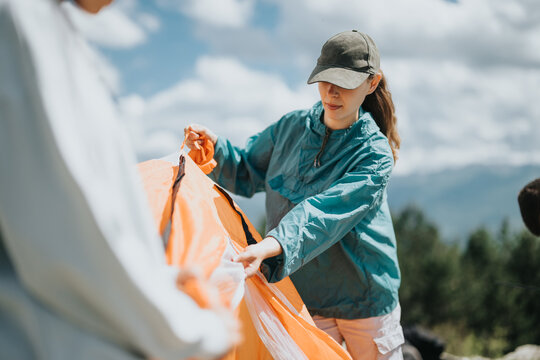 A young woman wearing a hat and jacket sets up an orange tent outdoors. The sky is filled with clouds, creating a serene setting.