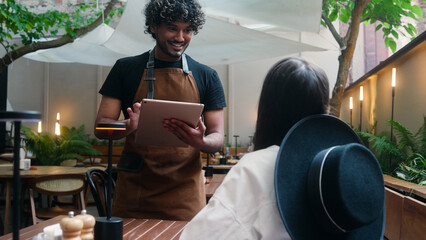 Friendly restaurant worker guy Arabian Indian male waiter in apron serve woman client in cafe...