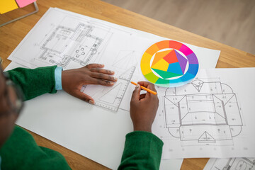 A person examines detailed architectural plans while using a color palette and drawing tools. The creative workspace is filled with sketches and planning materials, reflecting an artistic process.