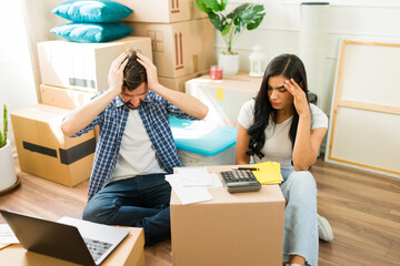 Young Hispanic couple surrounded by moving boxes in their new home, feeling overwhelmed while...