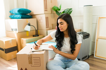 Latin woman sits on the floor surrounded by moving boxes, making a to-do list