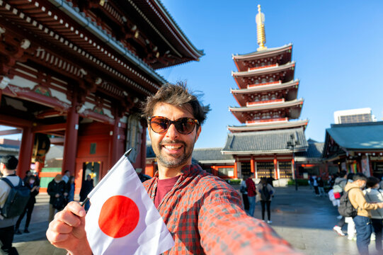 Handsome young tourist enjoying summer holiday in Tokyo, Japan - Traveling life style concept with smiling man taking selfie on city street with japan flag- Tourism and summertime vacation concept
