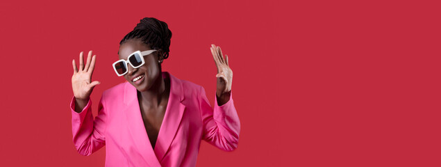 A black woman dressed in a bright pink blazer and oversized sunglasses is playfully posing with...