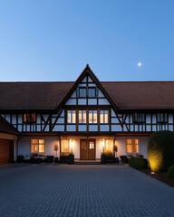 A Tudor-style house with warm glowing lights, surrounded by neatly trimmed hedges and a spacious cobblestone driveway under a clear evening sky with a bright celestial presence