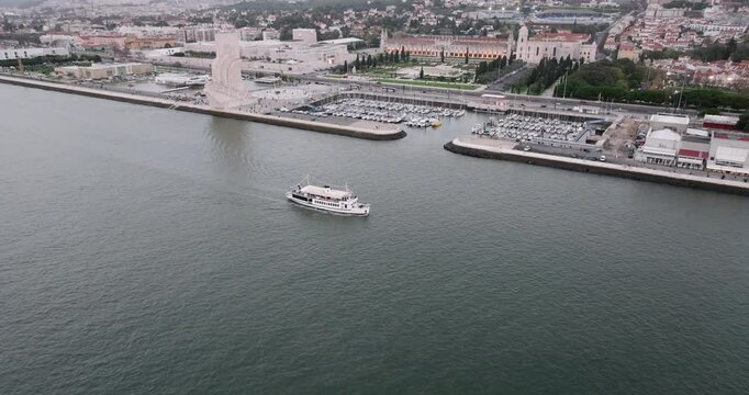 Aerial drone shot of motorboat passing by the Jeronimos Monastery and Discoveries monument on the waterfront in Lisbon, Portugal, Europe. Drone orbiting and descending. Shot in ProRes 422 HQ