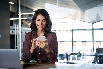 Young Indian business woman manager ceo using cell phone mobile app laptop. Smiling Latin Hispanic...