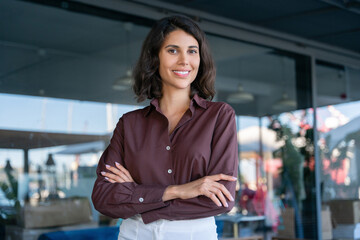 Portrait of beautiful successful hispanic young business woman with crossed arms smiling at camera. Latin or eastern confident middle age female ceo leader businesswoman standing in office. Copy space - obrazy, fototapety, plakaty