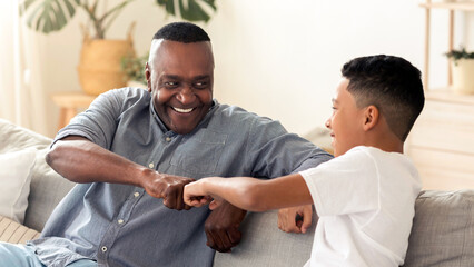 Fototapeta premium Male Family Unity. Happy African American Grandfather And Grandson Bumping Fists While Sitting On Couch At Home, Bonding Together