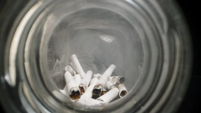 Dolly slider, close up. View through the mouth of a glass jar filled with cigarette butts, showing used filters and ash inside, emphasizing waste, pollution, and the impact of smoking in a contained s