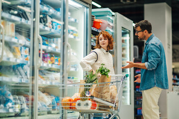 Couple discussing groceries shopping in supermarket refrigerator aisle