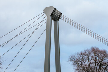 Obraz premium Close-up view of the structural cables and metal pillars of a modern suspension bridge against a cloudy sky. The cables are tensioned, connecting the tall steel supports.