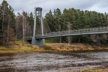 A low-angle view of a modern suspension bridge, showcasing the structural beams and cables. The bridge spans a natural landscape with tall evergreen trees and a cloudy sky in the background.