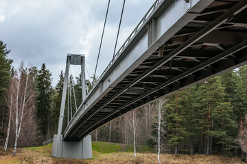 A low-angle view of a modern suspension bridge, showcasing the structural beams and cables. The bridge spans a natural landscape with tall evergreen trees and a cloudy sky in the background.