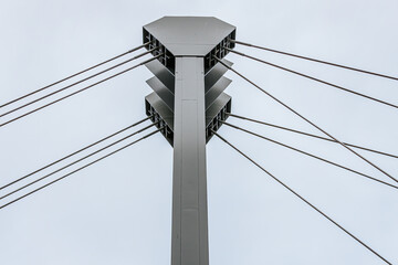 Close-up view of the structural cables and metal pillars of a modern suspension bridge against a cloudy sky. The cables are tensioned, connecting the tall steel supports.