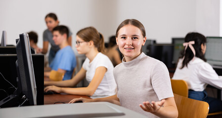 Fototapeta premium Cheerful teenage caucasian girl sitting at desk in computer class, explaining, gesturing and looking at camera.
