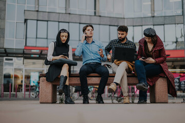 Fototapeta premium A group of young professionals using technology while sitting on a bench in a city setting. They are engaged in different activities, showcasing connectivity and modern work dynamics.