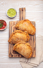 Three freshly baked empanadas with sesame seeds on a wooden board, white background, served with tomato salsa and guacamole. Traditional dish of Spain and Latin America