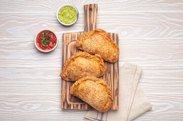 Three freshly baked empanadas with sesame seeds on a wooden board, white background, served with tomato salsa and guacamole. Traditional dish of Spain and Latin America