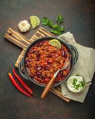 Traditional mexican chili con carne served in a cast iron pot, garnished with lime and red chili peppers, alongside sour cream and fresh ingredients on a rustic wooden board