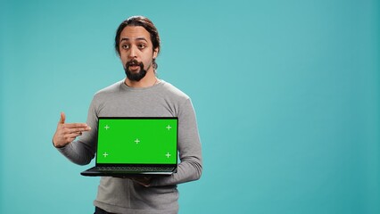 Man presenting green screen mobile laptop, isolated over studio background. Arab person holding copy space chroma key notebook device used for advertising brands, camera B