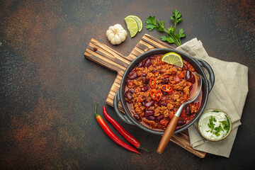 Traditional mexican chili con carne served in a cast iron pot, garnished with lime and red chili peppers, alongside sour cream and fresh ingredients on a rustic wooden board, copy space