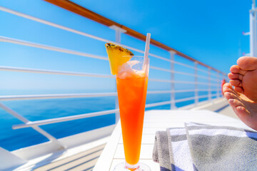 A female cruise ship passenger relaxes with her feet up and a tropical cocktail drink with fresh pineapple, on the upper deck of a large cruise ship at sea.