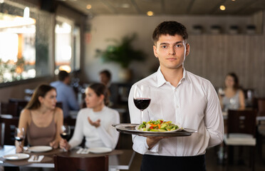 Young male waiter posing with tray of salad and wine in restaurant