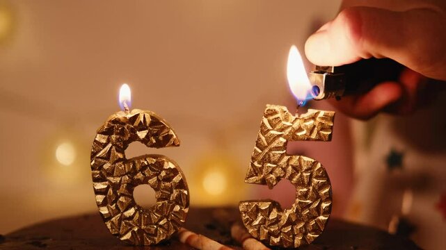 Happy Birthday Chocolate Cake with golden 65 Candles indoor with garland. A human hand lights candles using a lighter in a cozy home festive atmosphere. Close up view
