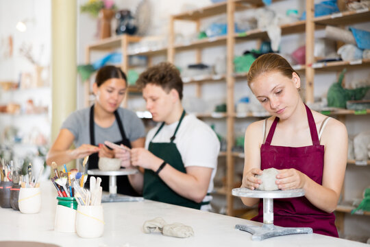 Young female teacher in apron helps teenager boy and teenager girl students to make product from clay in ceramic workshop